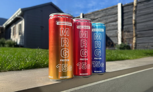 MRG Protein cans all flavours Blue Razberry, Orange Crush and Pink Límonada lined up in a sunny outside setting with grass and blue sky.